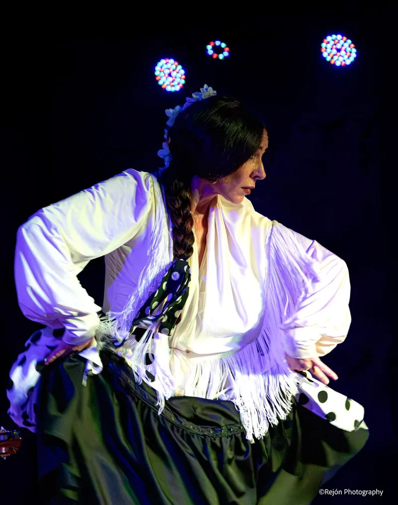 Bailaora en primer plano con camisa blanca de flecos y mantón de lunares, con los focos de colores del techo al fondo