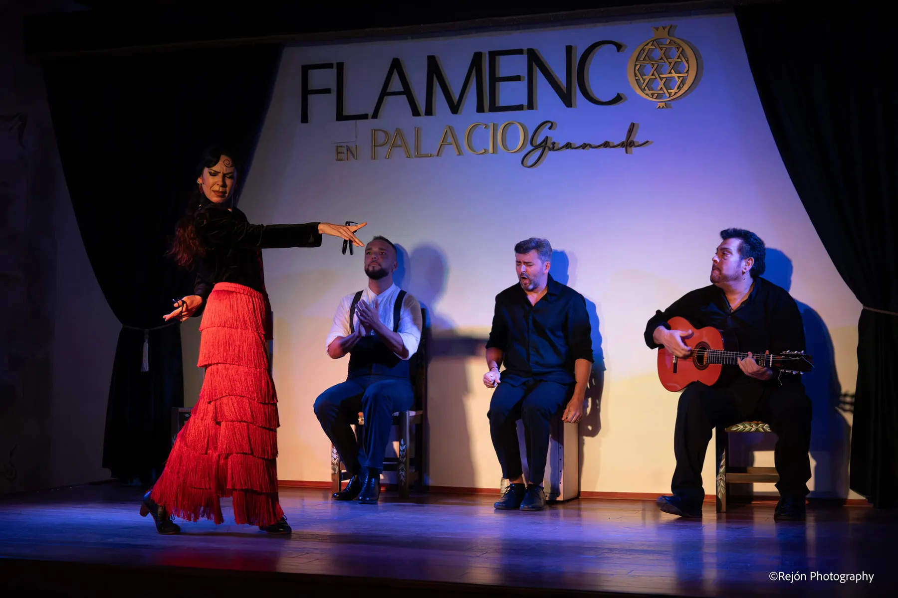 Bailaora con falda roja de flecos y castañuelas junto al palmero, el cantaor y el guitarrista, sentados en fila sobre el escenario
