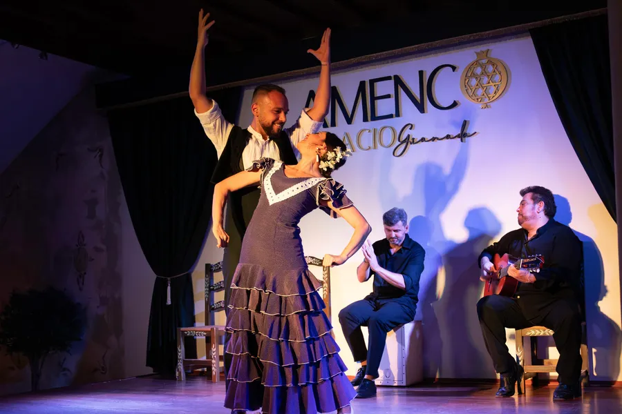 Bailaora arqueada hacia atrás con traje de volantes y una flor en el pelo, un bailaor con los brazos en alto detrás de ella, y el palmero y el guitarrista sentados al fondo