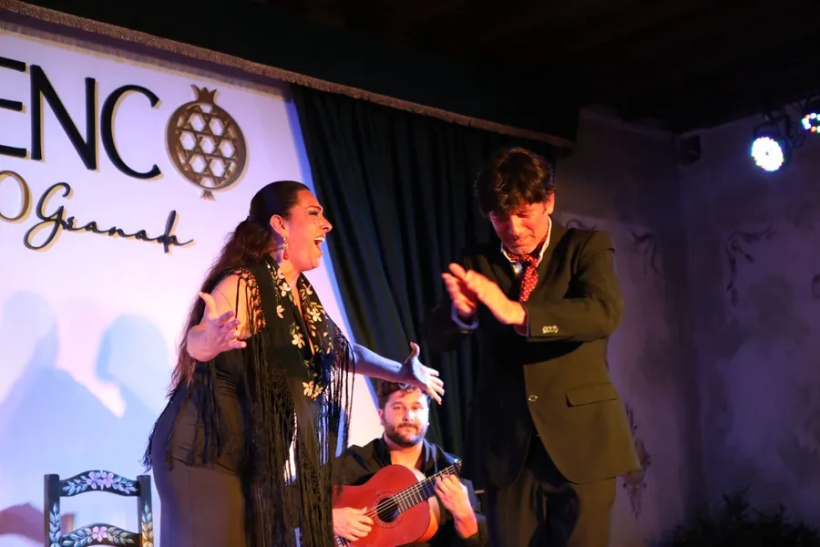 Cantaora con mantón de flecos cantando con los brazos abiertos, un bailaor marcándole las palmas y el guitarrista tocando sentado detrás