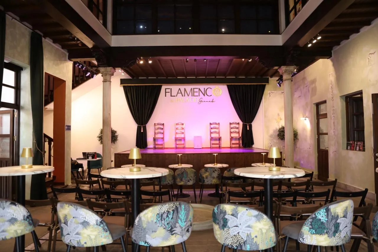 Sala del tablao vista desde el fondo, con el escenario iluminado en violeta, las sillas de enea sobre las tablas y las mesas de mármol con lámparas doradas